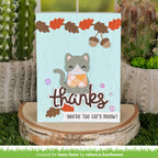 A handmade autumn-themed greeting card featuring a cute gray cat with an orange patterned sweater, acorns, and the words 'thanks' and 'you're the cat's meow!' displayed on a rustic wood slice stand, with cacti in the background.
