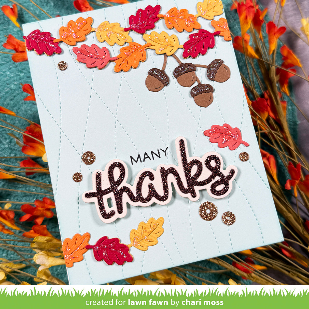 A handmade autumn-themed greeting card with 'MANY thanks' in raised glittery lettering, decorated with layered die-cut fall leaves and acorns. The card is displayed on a textured teal surface surrounded by dried orange and red leaves. | color-code:alt1