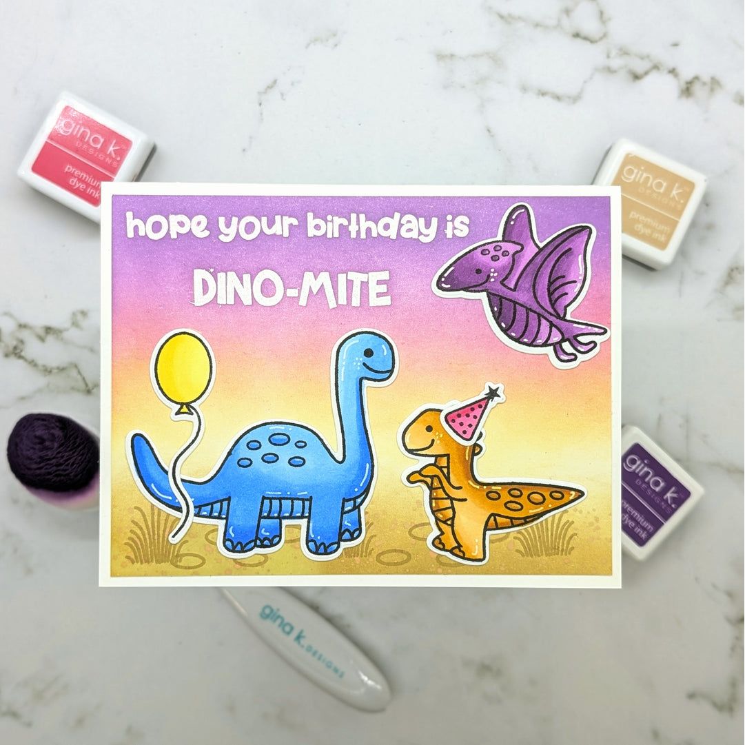 A handmade birthday card featuring cartoon dinosaurs and the text 'hope your birthday is DINO-MITE' on a pastel gradient background, styled on a white marble surface with Gina K. ink pads and a blending brush nearby.