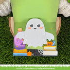 A handmade card featuring a cute white ghost character with pink cheeks, standing on a green grass-like surface. The cover shows a purple potion bottle, books, a pumpkin, spiderweb, and stars, with a green backing board behind the craft.