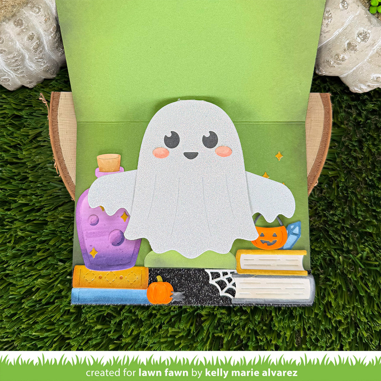 A handmade card featuring a cute white ghost character with pink cheeks, standing on a green grass-like surface. The cover shows a purple potion bottle, books, a pumpkin, spiderweb, and stars, with a green backing board behind the craft.