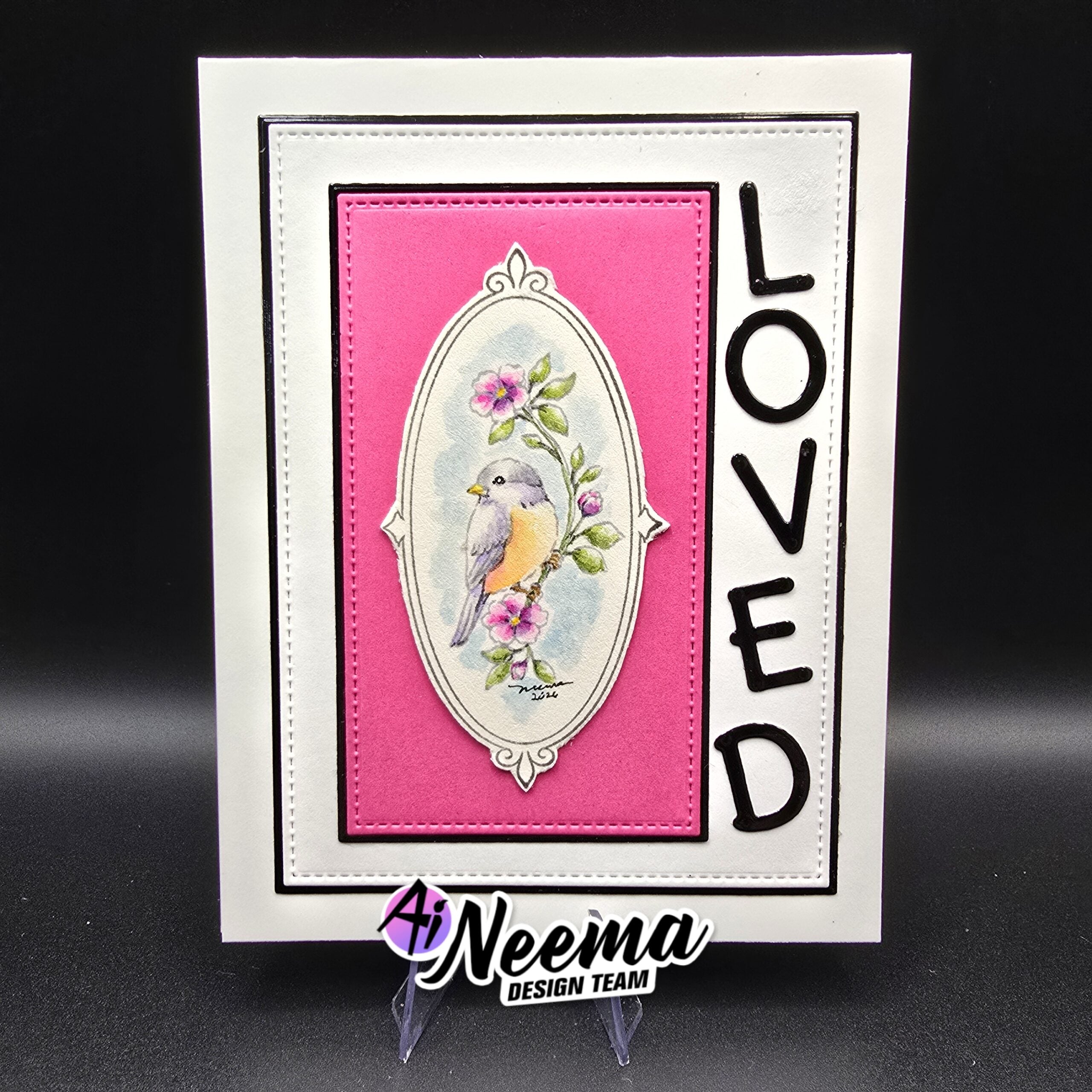 A handmade greeting card displayed on a black background, featuring a white border, a bright pink inner panel, an oval illustration of a small bird with pink flowers, and the word 'LOVED' printed vertically in black on the right. 