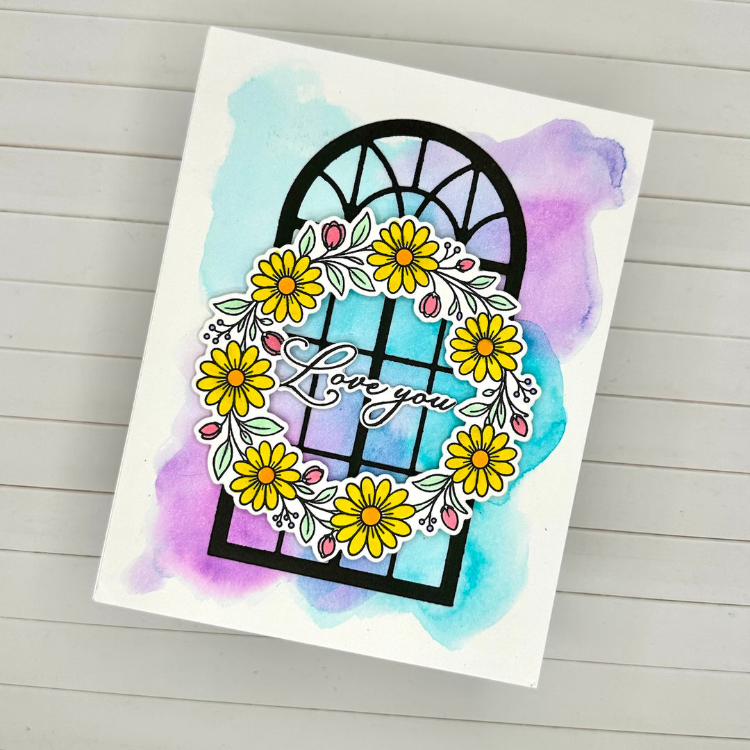 A handmade greeting card featuring a black arched window outline surrounded by yellow daisies, green leaves, and pink accents, with the words 'Love you' in script over a blue and purple watercolor wash. The card is photographed on a light gray wood plank surface.