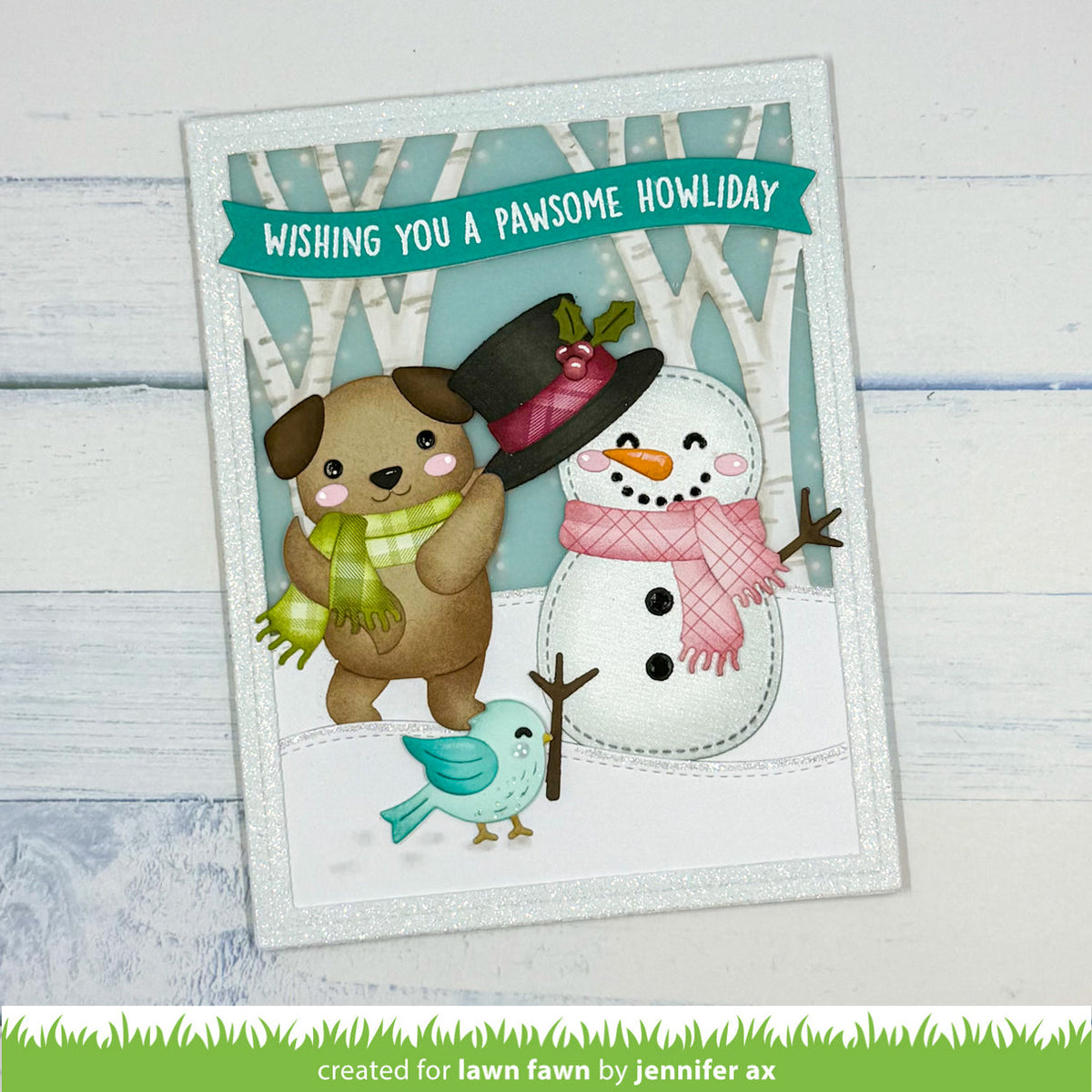 A handmade greeting card featuring a brown puppy in a green scarf, a snowman with a pink scarf and black hat, and a small blue bird, with the text 'WISHING YOU A PANSOME HOWIDAY' on a teal banner. The card is displayed on a white wood background.