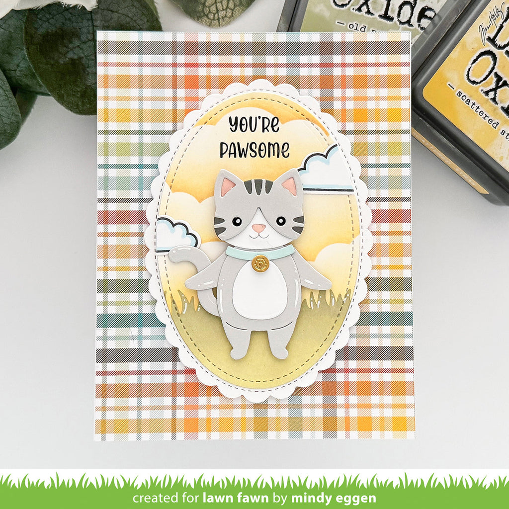 A handmade greeting card featuring a cute gray tabby cat layered over a yellow oval with the text 'YOU'RE PAWSOME'. The card has a multicolor plaid background in orange, yellow, green, and white, and is displayed on a white surface with craft ink pads in the background. | color-code:alt1