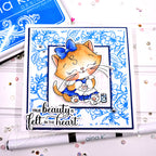 A handmade greeting card featuring a cute orange cartoon cat in a blue bow and blue dress, surrounded by blue floral patterns. The card has the text 'TRUE beauty is felt in the heart' and is displayed on a white surface with craft tools and rhinestones.