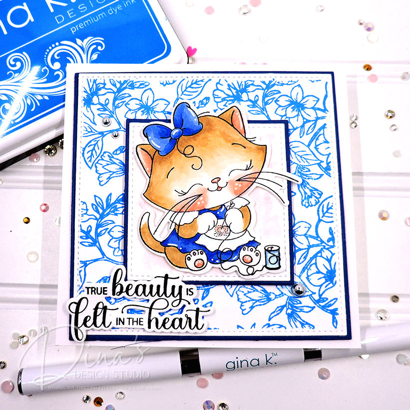 A handmade greeting card featuring a cute orange cartoon cat in a blue bow and blue dress, surrounded by blue floral patterns. The card has the text 'TRUE beauty is felt in the heart' and is displayed on a white surface with craft tools and rhinestones.
