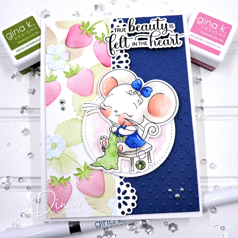 A handmade greeting card featuring a cute white mouse in a blue dress and bow, sitting on a stool and holding a green bag. The card has a navy blue textured background on the right, a strawberry and white flower patterned background on the left, and the phrase 'TRUE beauty is felt IN THE heart' in white script.