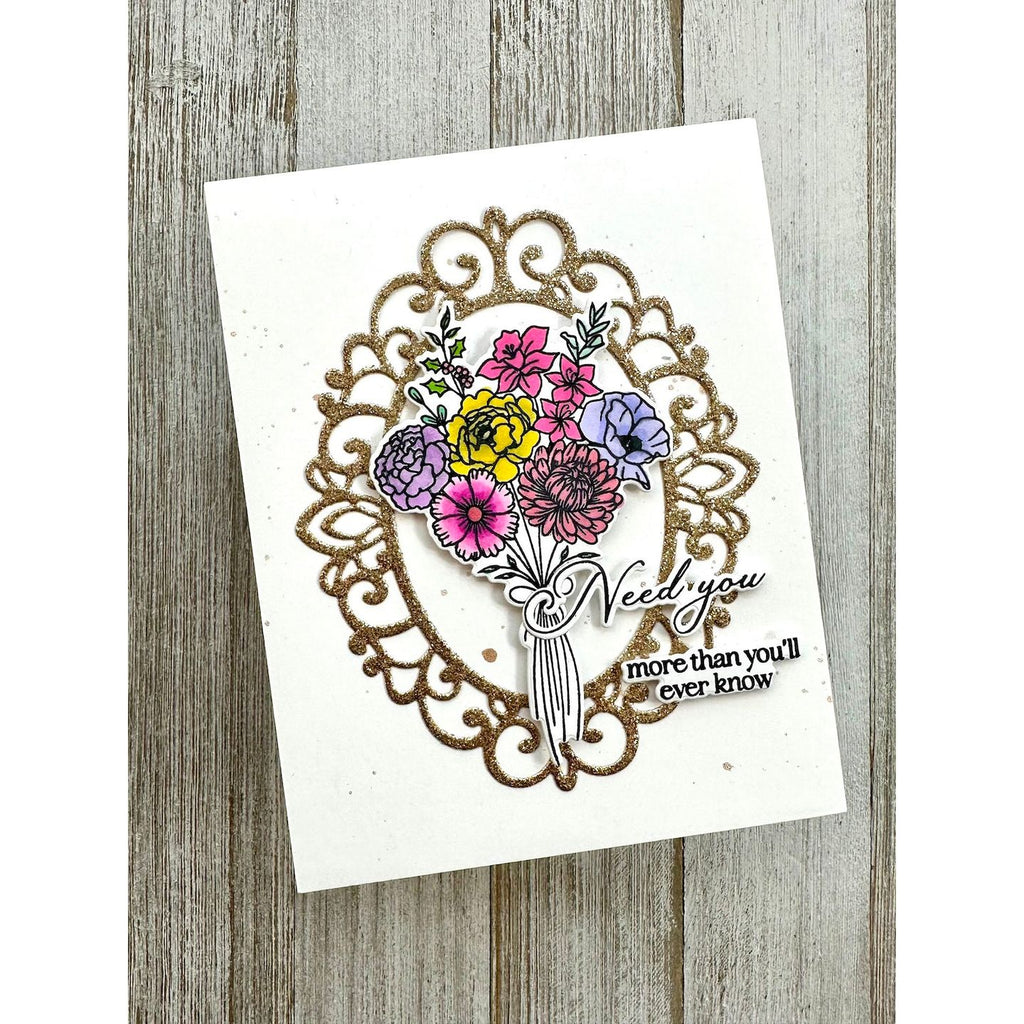 A handmade greeting card featuring a glittery gold ornate frame, a colorful floral bouquet with yellow, pink, purple, and blue flowers, and the words 'Need you more than you'll ever know' on a white card, displayed on a rustic gray wood background.