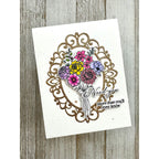A handmade greeting card featuring a glittery gold ornate frame, a colorful floral bouquet with yellow, pink, purple, and blue flowers, and the words 'Need you more than you'll ever know' on a white card, displayed on a rustic gray wood background.