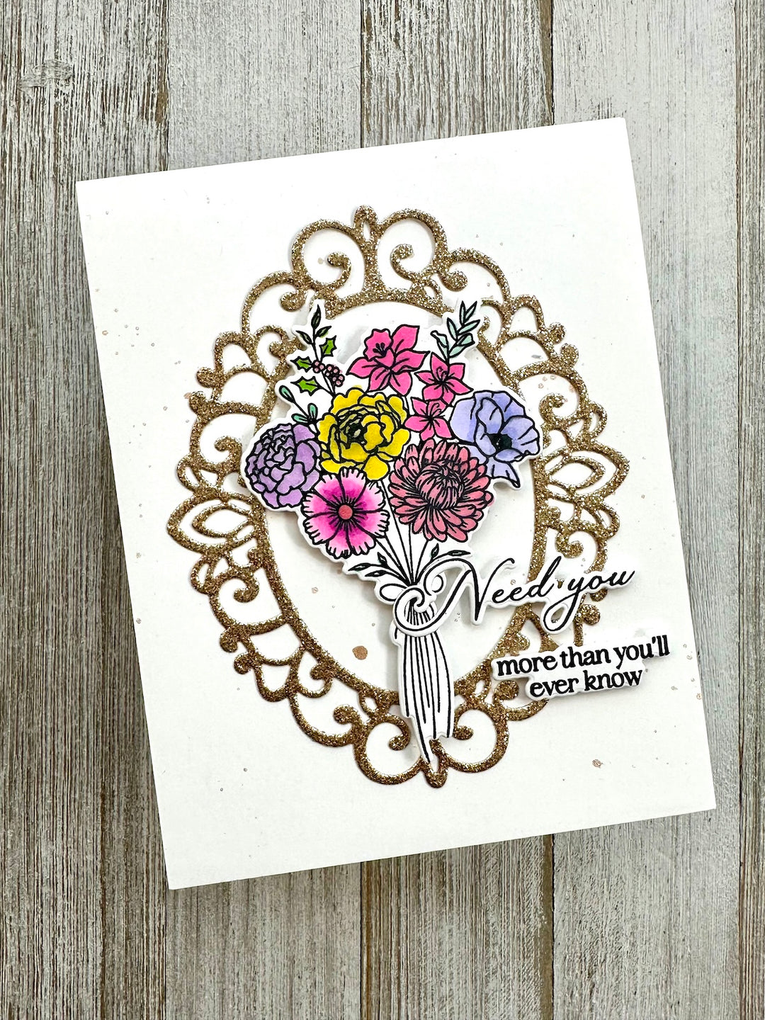 A handmade greeting card featuring a glittery gold ornate frame, a colorful floral bouquet with yellow, pink, purple, and blue flowers, and the words 'Need you more than you'll ever know' on a white card, displayed on a rustic gray wood background.