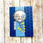 A handmade greeting card featuring a layered blue and navy design with polka dots, textured stripes, and a central circular motif with orange and yellow botanical elements, displayed on a rustic wooden background.