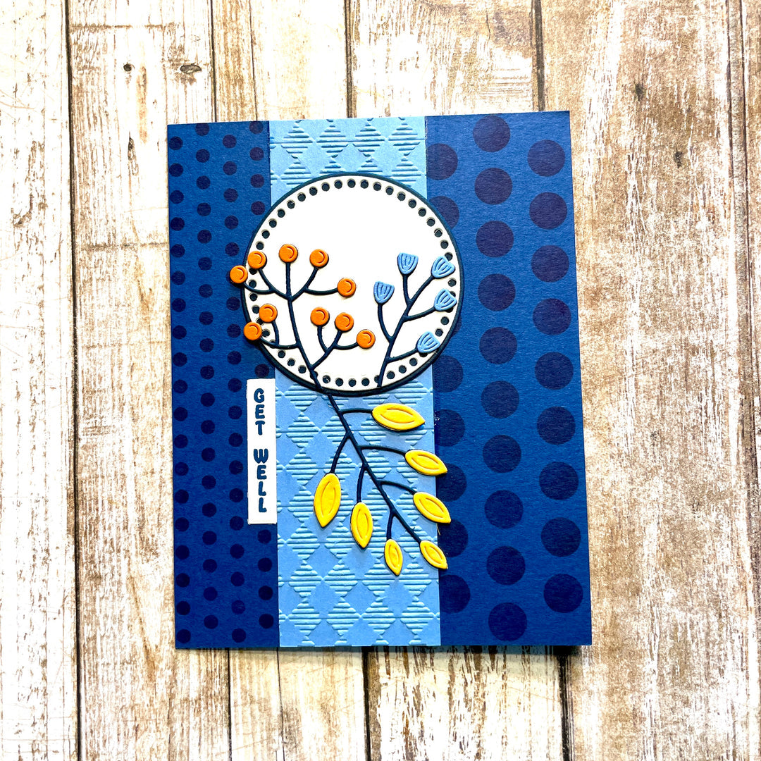A handmade greeting card featuring a layered blue and navy design with polka dots, textured stripes, and a central circular motif with orange and yellow botanical elements, displayed on a rustic wooden background.