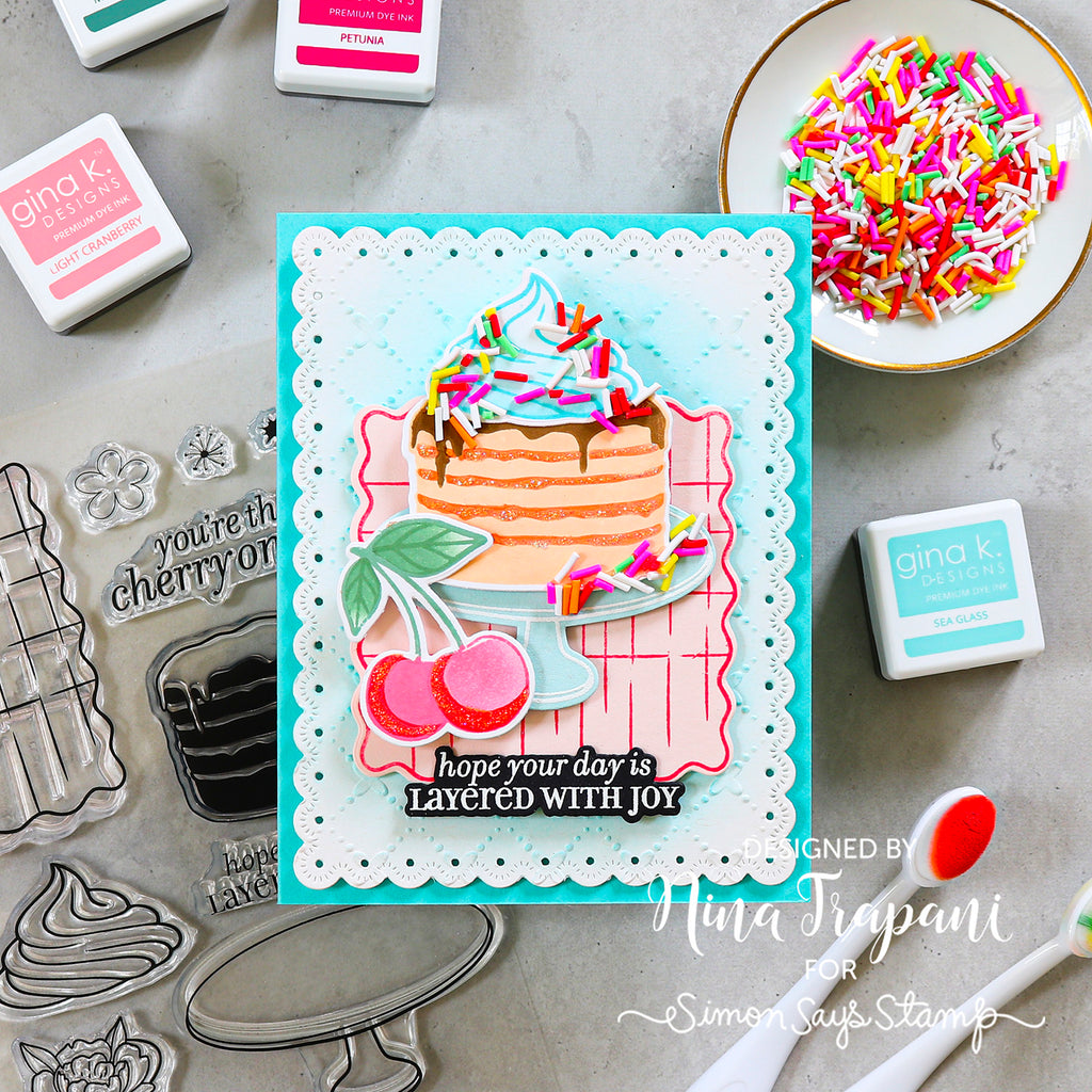 A handmade greeting card featuring a layered cake with sprinkles, cherries, and the text 'hope your day is LAYERED WITH JOY', styled in a flat-lay with Gina K. stamps, ink pads, and sprinkles on a gray surface.