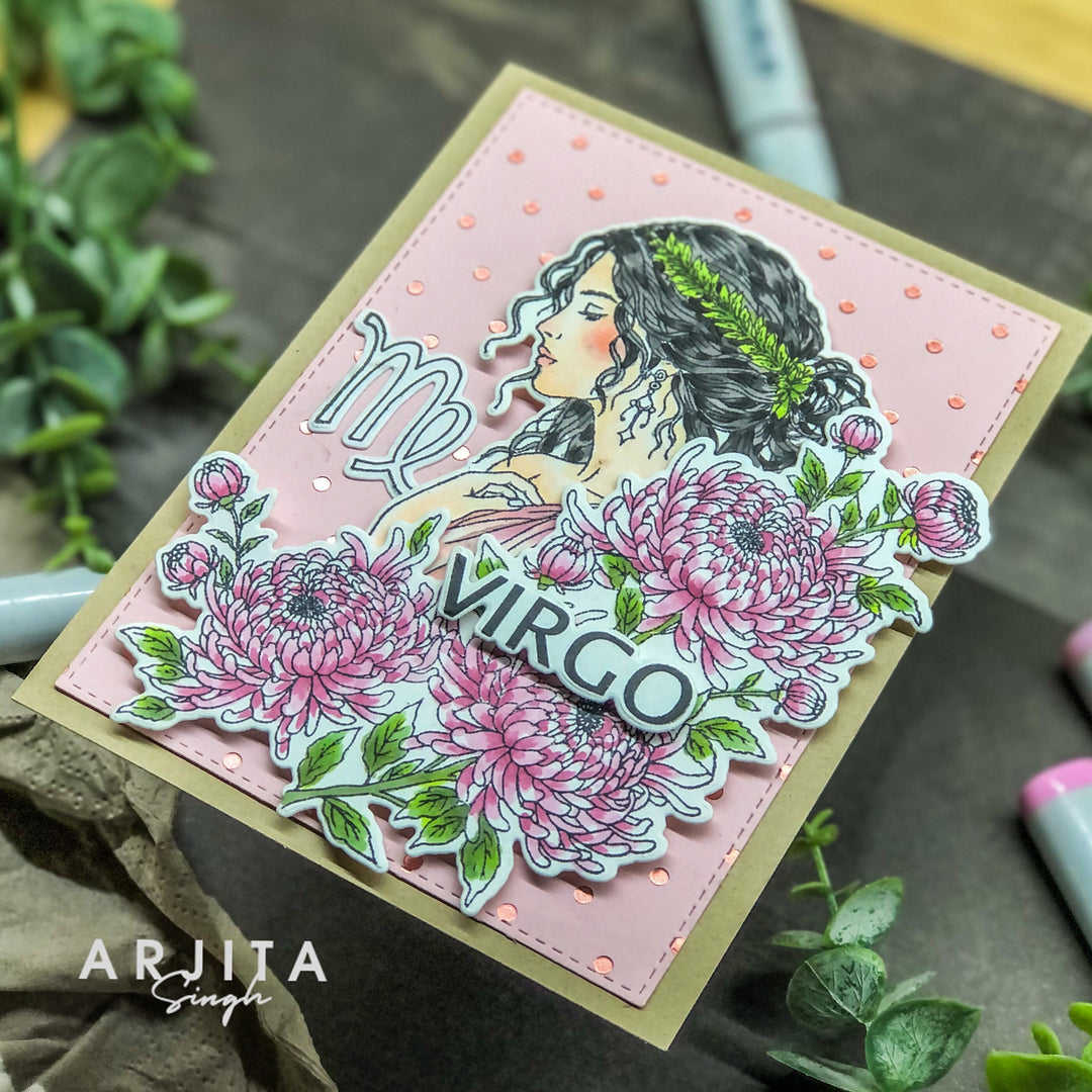 A handmade greeting card featuring a pink background with a die-cut illustration of a woman, pink flowers, green leaves, and the word 'VIRGO' with a star. The card is placed on a dark surface with decorative plants and markers around it.