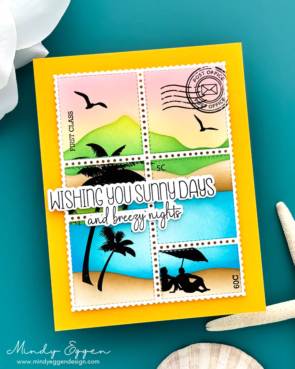 A handmade greeting card featuring a postage stamp collage design with beach scenes and the text 'WISHING YOU SUNNY DAYS and breezy nights,' photographed on a teal surface with decorative seashells and a white flower.