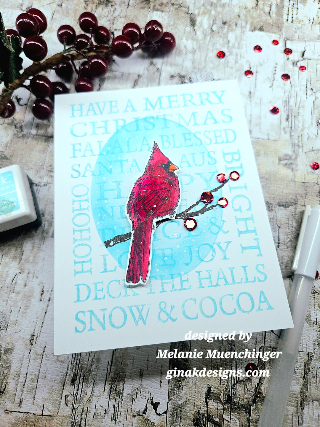 A handmade greeting card featuring a red cardinal perched on a branch with red berries, set against a light blue circular background and a white card with festive text. The card is styled on a rustic wooden surface with holiday decorations.