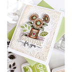 A handmade greeting card featuring a stamped and embossed floral design with sunflowers in a pot and the text 'GRATEFUL for you'. The card is styled on top of green and white cardstock with decorative paper flowers and ink pads around it.