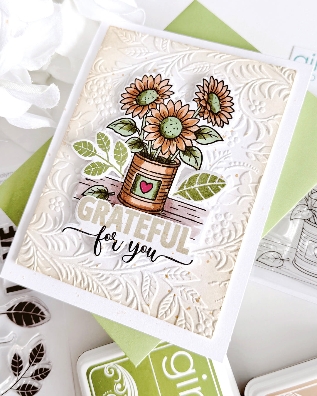 A handmade greeting card featuring a stamped and embossed floral design with sunflowers in a pot and the text 'GRATEFUL for you'. The card is styled on top of green and white cardstock with decorative paper flowers and ink pads around it.