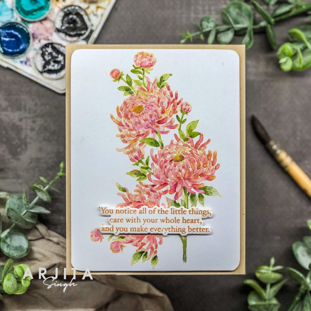 A handmade greeting card featuring a watercolor-style floral design with pink and orange chrysanthemums and a layered quote, styled on a dark textured surface with greenery, a paint palette, and a paintbrush.