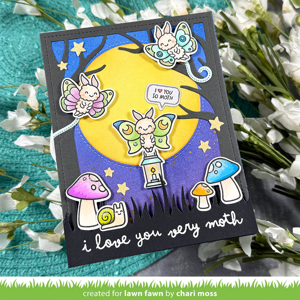 A handmade greeting card featuring a whimsical moth-themed illustration, displayed on a textured teal surface with white flowers. The card has a black border and shows three colorful butterflies, a large yellow moon, a snail under a purple mushroom, and the text 'i love you very moth'. | color-code:alt1