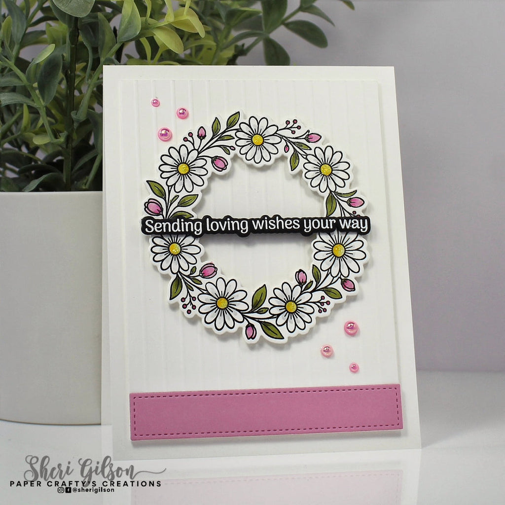 A handmade greeting card featuring a white daisy wreath with green leaves, pink buds, and a black banner that reads 'Sending loving wishes your way.' The card has a pink rectangular accent at the bottom and is photographed standing upright on a white surface with a potted plant in the background.