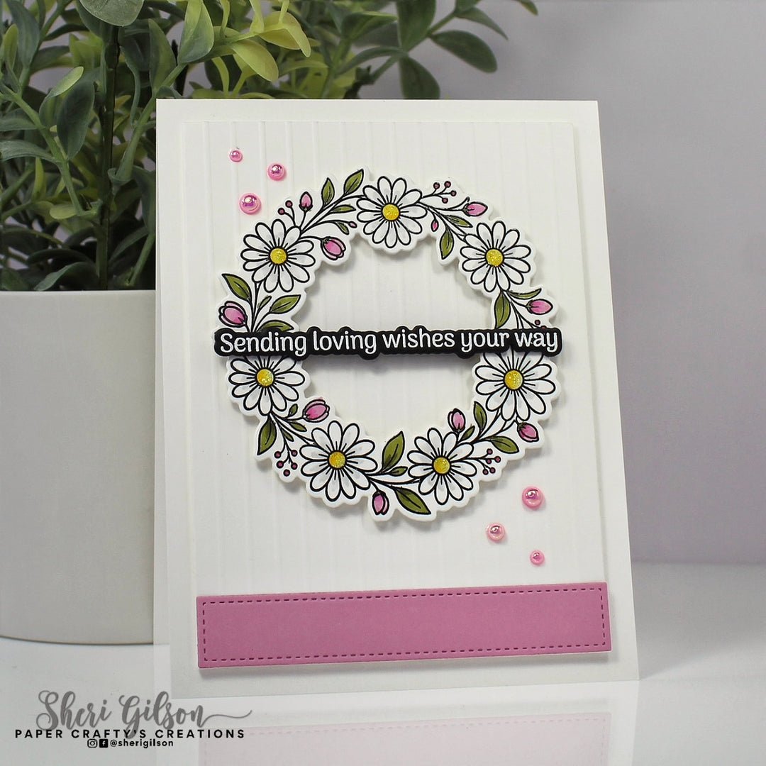 A handmade greeting card featuring a white daisy wreath with green leaves, pink buds, and a black banner that reads 'Sending loving wishes your way.' The card has a pink rectangular accent at the bottom and is photographed standing upright on a white surface with a potted plant in the background.