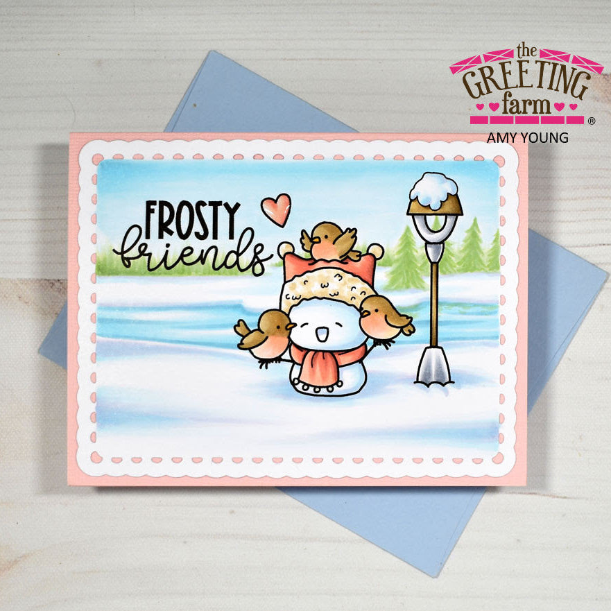 A handmade greeting card featuring a winter scene with a snowman wearing a hat and scarf, surrounded by small birds, with the text 'FROSTY friends'. The card is displayed on top of a blue envelope on a light wood background, with branding for 'The Greeting Farm' visible.