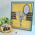 A handmade greeting card featuring a yellow embossed background, a bouquet of white daisies with yellow centers, brown lace strips, and a white oval label with a Bible verse. The card is displayed upright next to a round yellow bee-themed pin, with a white background and a watermark at the bottom.