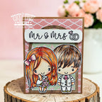 A handmade greeting card featuring cartoon-style bride and groom characters with the text 'Mr & Mrs' and a ring icon, displayed on a rustic wood slice with a soft pink floral background. The greeting card is branded with 'The Greeting Farm' and 'Cynthia Roybal' text.