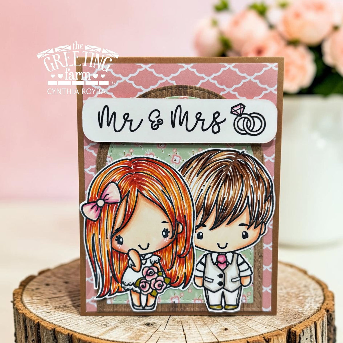 A handmade greeting card featuring cartoon-style bride and groom characters with the text 'Mr & Mrs' and a ring icon, displayed on a rustic wood slice with a soft pink floral background. The greeting card is branded with 'The Greeting Farm' and 'Cynthia Roybal' text.