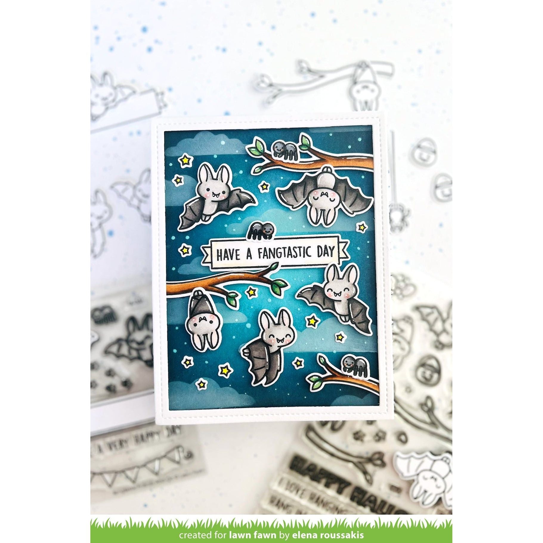 A handmade greeting card featuring cute cartoon bats flying on branches against a blue starry night sky, with the text 'HAVE A FANTASTIC DAY'. The card is displayed on a white surface with clear stamp designs in the background.
