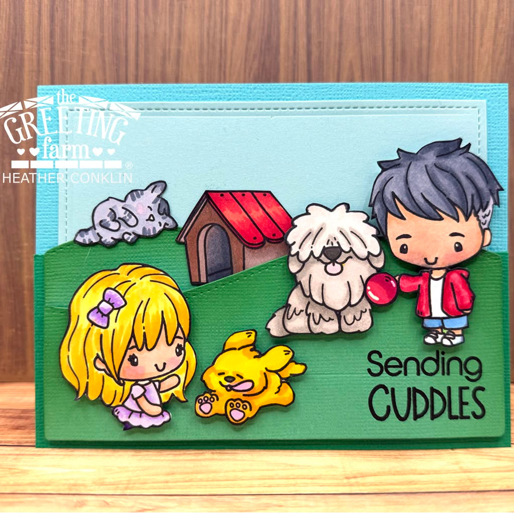 A handmade greeting card featuring cute chibi-style cartoon children, two dogs, a dog house, and the text 'Sending CUDDLES' on a green and light blue layered card, photographed on a wooden surface.
