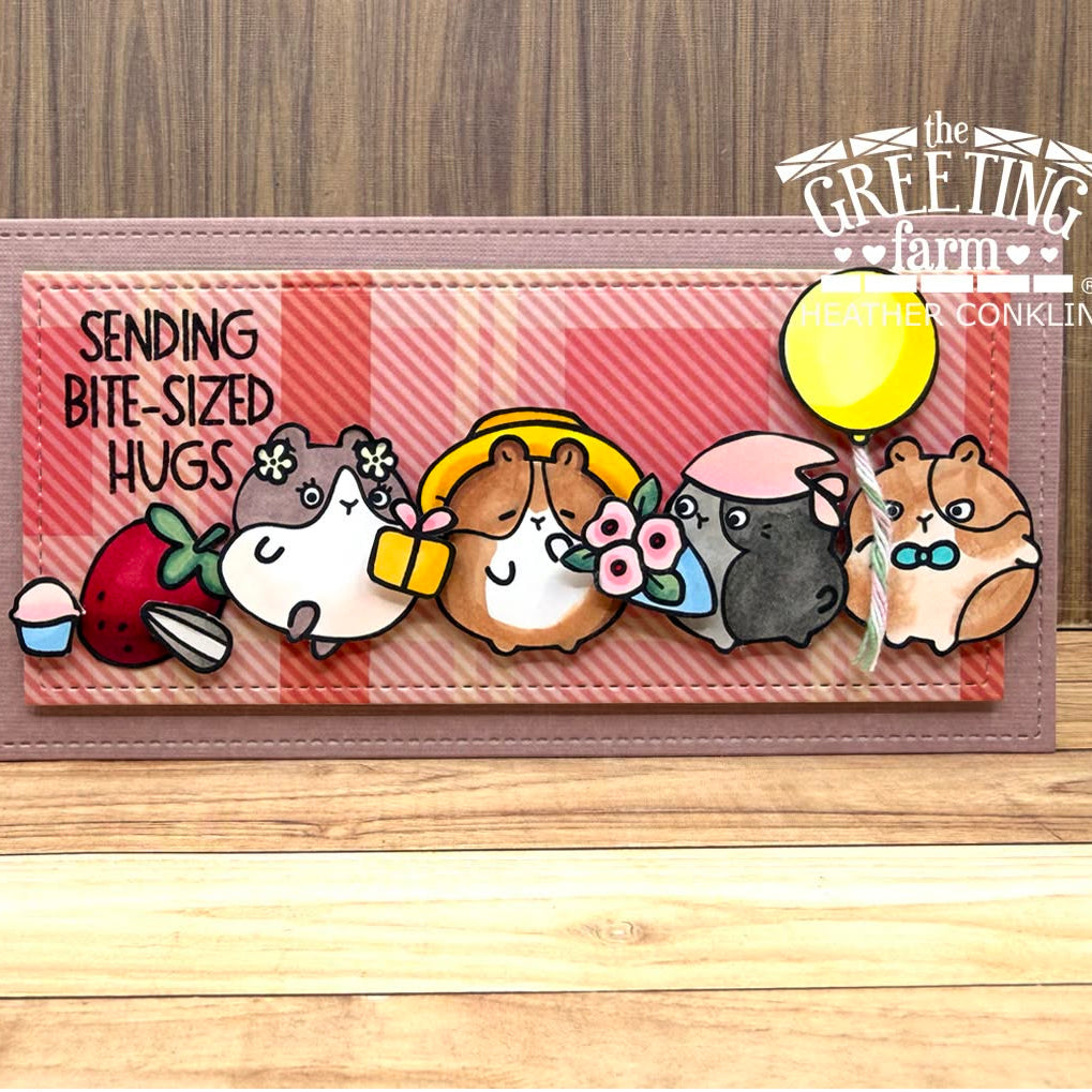 A handmade greeting card featuring cute hamster illustrations and the text 'SENDING BITE-SIZED HUGS' on a pink plaid background, displayed on a wooden surface with a wood-grain backdrop. The card has a stitched mauve border and includes a yellow balloon, strawberry, cupcake, and hamsters in different colors.