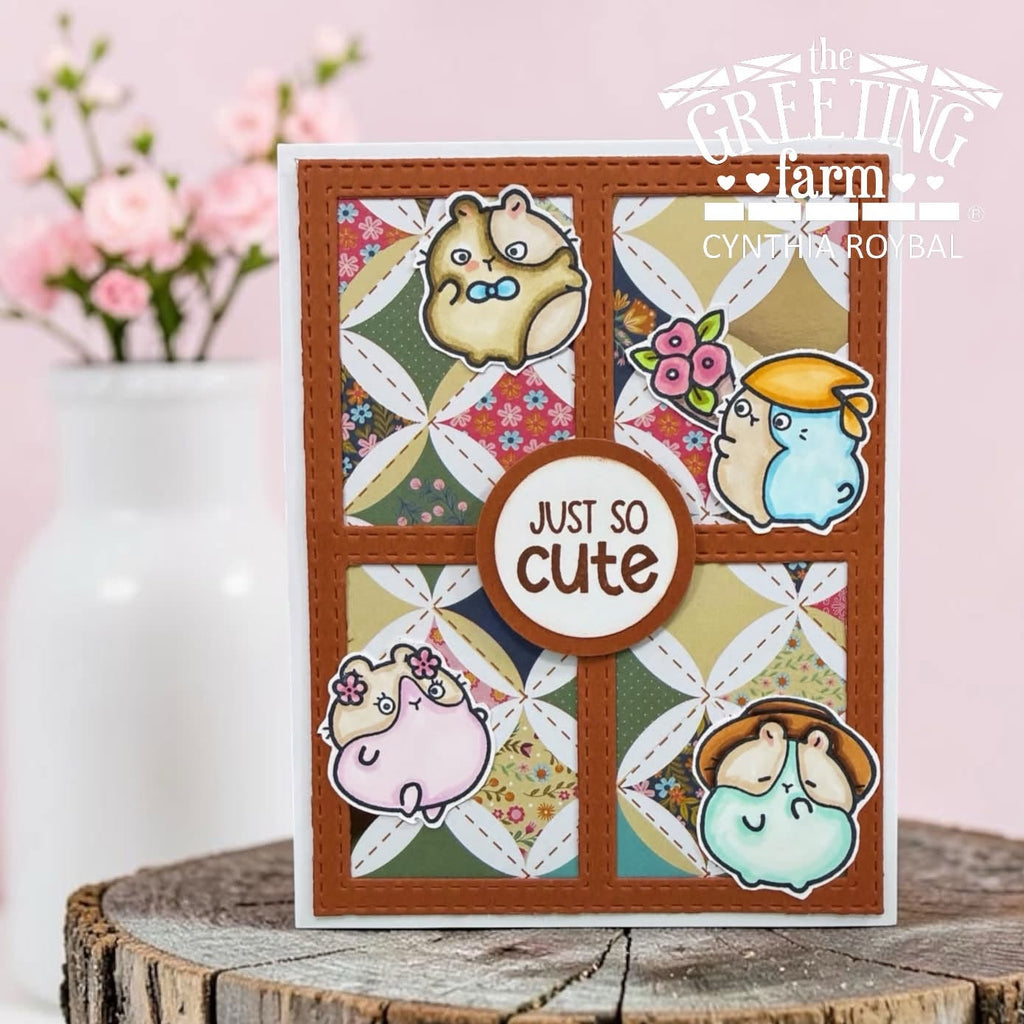 A handmade greeting card featuring four cute hamster illustrations arranged in a quilt pattern with a central round label that reads 'JUST SO cute'. The card is displayed on a rustic wood slice with a soft-focus pink floral background.