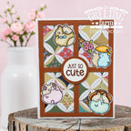 A handmade greeting card featuring four cute hamster illustrations arranged in a quilt pattern with a central round label that reads 'JUST SO cute'. The card is displayed on a rustic wood slice with a soft-focus pink floral background.