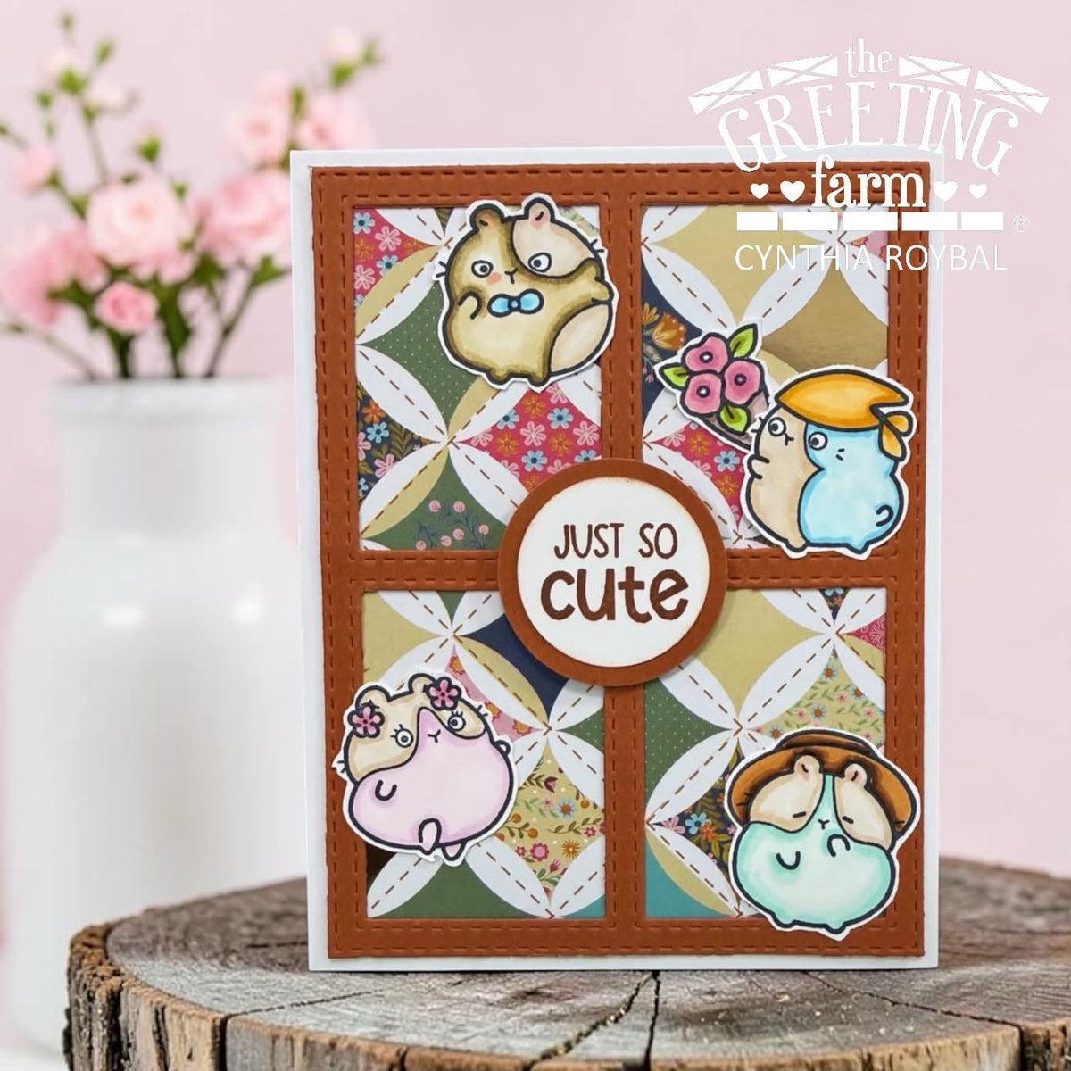 A handmade greeting card featuring four cute hamster illustrations arranged in a quilt pattern with a central round label that reads 'JUST SO cute'. The card is displayed on a rustic wood slice with a soft-focus pink floral background.
