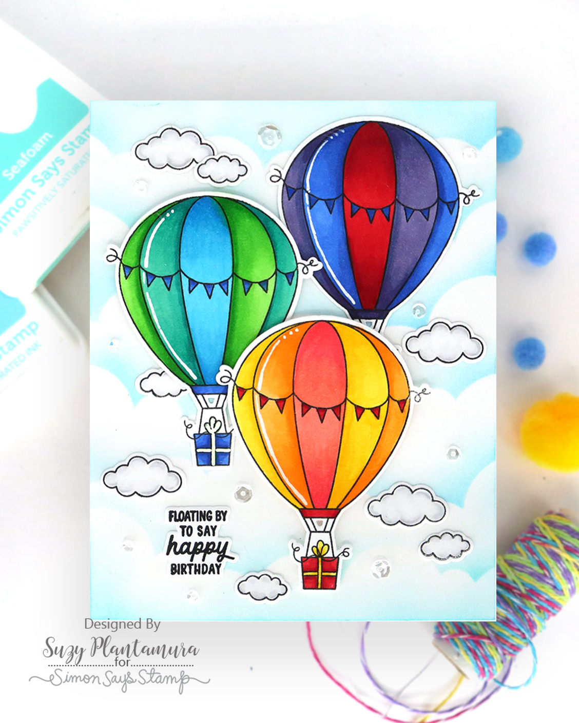 A handmade greeting card featuring three colorful hand-painted hot air balloons, clouds, and the text 'FLOATING BY TO SAY happy birthday'. The card is displayed on a white surface with craft supplies Simon Says Clear Stamps Above the Clouds 1498ssc Suzy Plantamura | color-code:ALT01