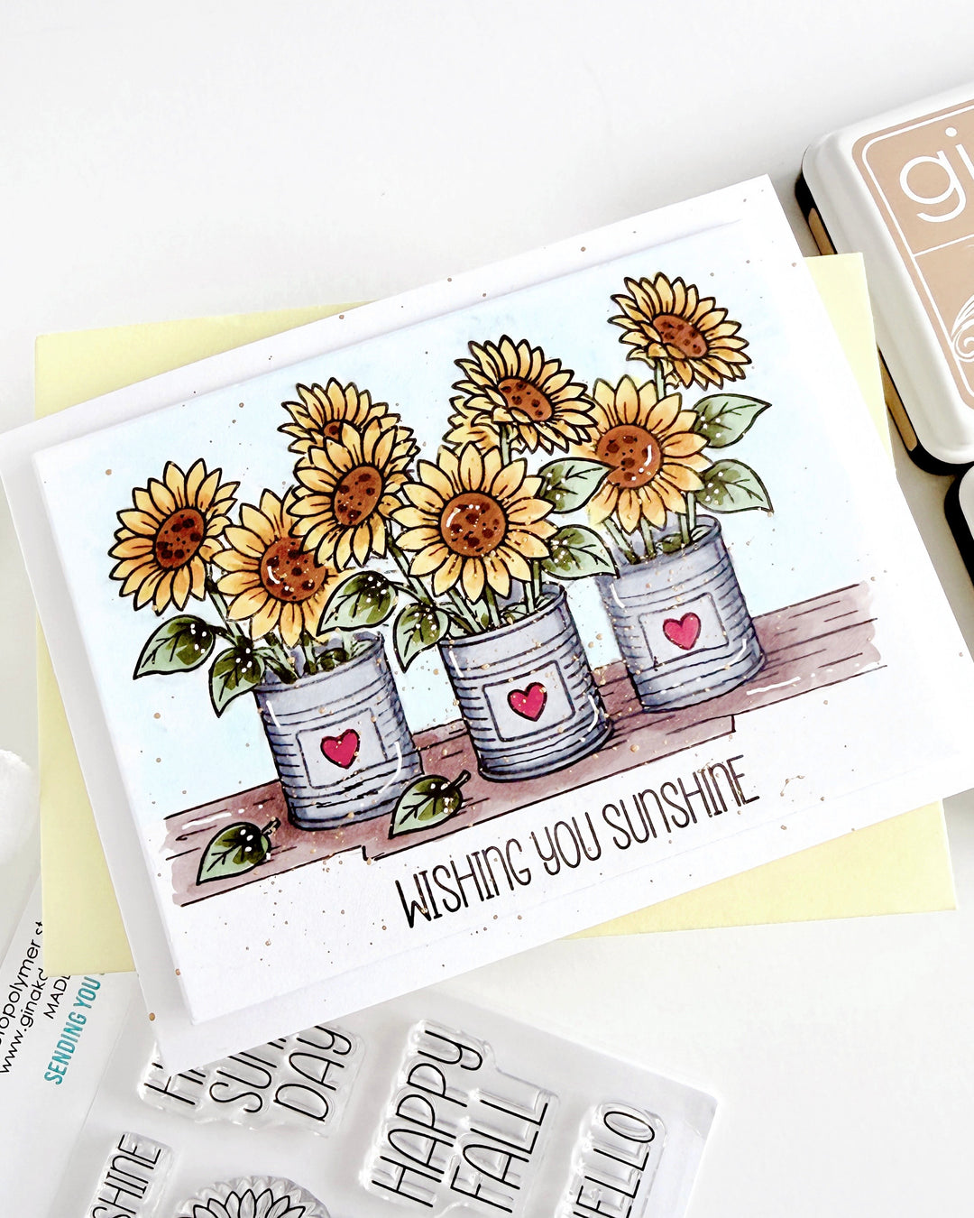 A handmade greeting card featuring three potted sunflowers with the text 'WISHING YOU SUNSHINE' on a white background, styled with crafting tools and stamps.