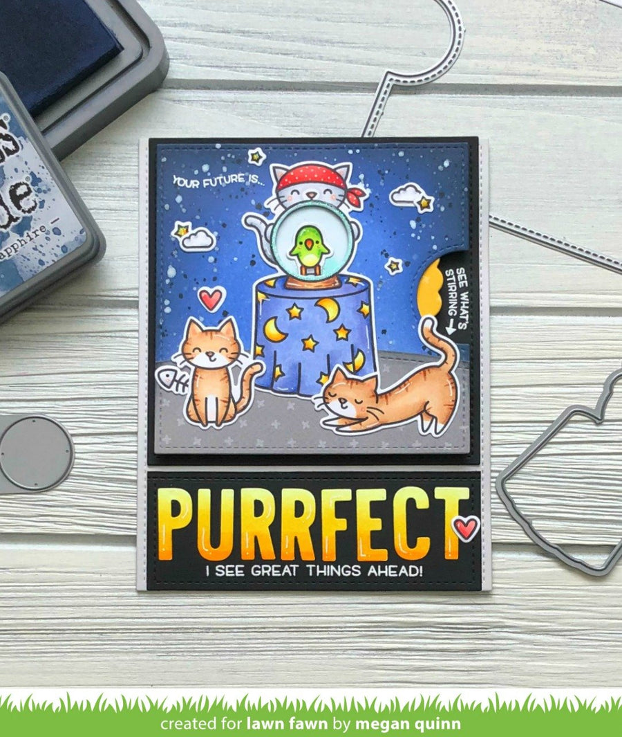 A handmade greeting card featuring two illustrated cats and a whimsical space-themed design, displayed on a light wood surface with crafting tools partially visible around it.