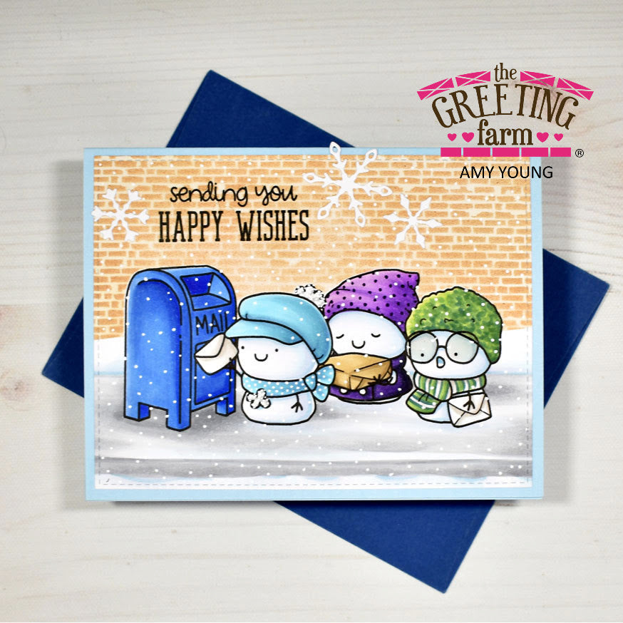 A handmade greeting card from The Greeting Farm by Amy Young, featuring three cute winter cartoon characters by a blue mailbox on a snowy ground, with a brick wall background and the text 'sending you HAPPY WISHES'. The card is displayed on top of a dark blue envelope on a light wood background.