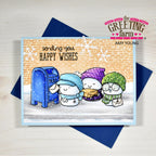 A handmade greeting card from The Greeting Farm by Amy Young, featuring three cute winter cartoon characters by a blue mailbox on a snowy ground, with a brick wall background and the text 'sending you HAPPY WISHES'. The card is displayed on top of a dark blue envelope on a light wood background.