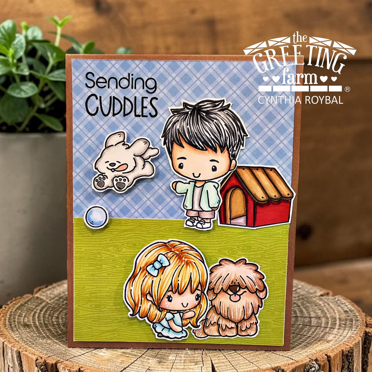 A handmade greeting card with a brown border, featuring cartoon-style children and dogs on a blue plaid and olive green background. The card is displayed on a rustic wood slice with a potted plant in the background.