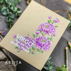 A handmade greeting card with a kraft paper background, featuring purple chrysanthemum illustrations and the text 'VIRGO You are the perfect friend!'. The card is photographed at an angle on a dark textured surface with green leaves and colored pencils as props.