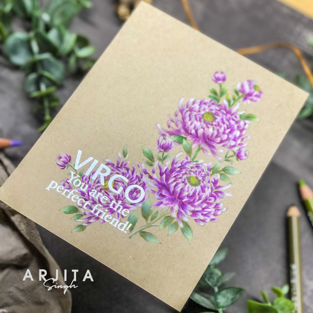 A handmade greeting card with a kraft paper background, featuring purple chrysanthemum illustrations and the text 'VIRGO You are the perfect friend!'. The card is photographed at an angle on a dark textured surface with green leaves and colored pencils as props.