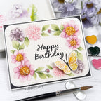 A handmade greeting card with a watercolor floral design and a butterfly, reading 'Happy Birthday' in black script. The card is styled on a white surface with a paintbrush, heart-shaped paint palettes, and blurred flowers in the background.