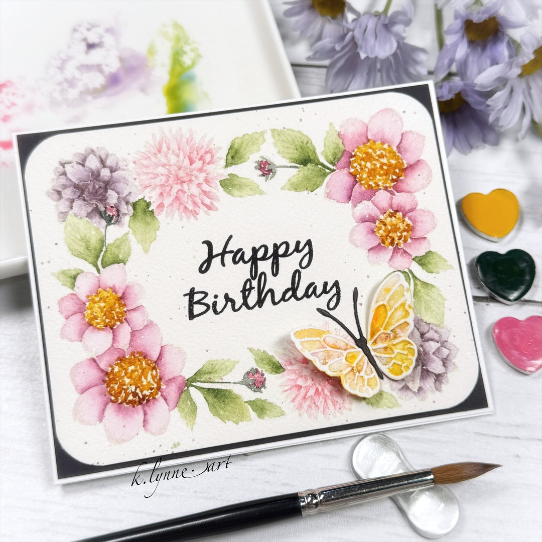 A handmade greeting card with a watercolor floral design and a butterfly, reading 'Happy Birthday' in black script. The card is styled on a white surface with a paintbrush, heart-shaped paint palettes, and blurred flowers in the background.