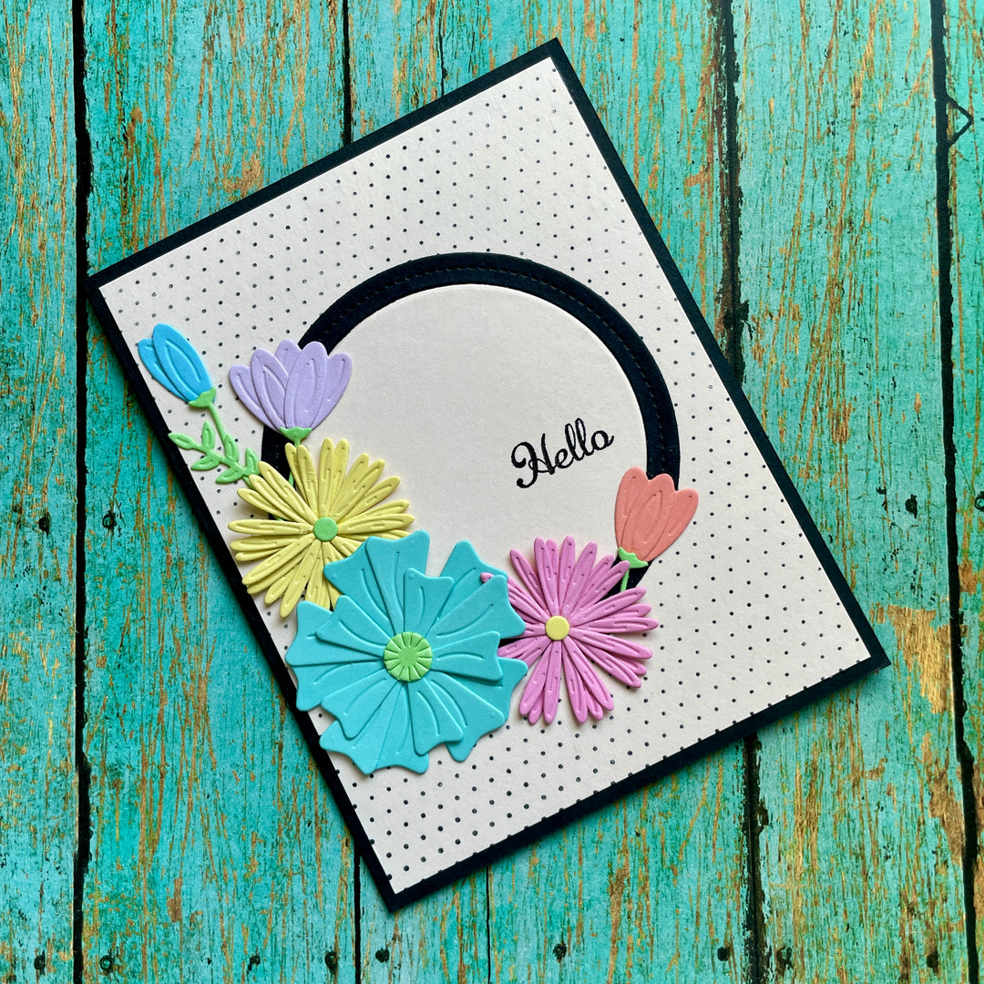 A handmade greeting card with a white polka-dot background, a black border, a black arch, and the word 'Hello' in script. The card is decorated with layered paper flowers in blue, yellow, pink, and purple, and is photographed on a distressed turquoise wooden surface.