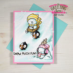 A handmade greeting card with a winter cartoon scene, shown with a teal envelope on a light wood background. The card features three cute penguins in winter clothing, including one sliding in a sled and another wearing pink earmuffs.