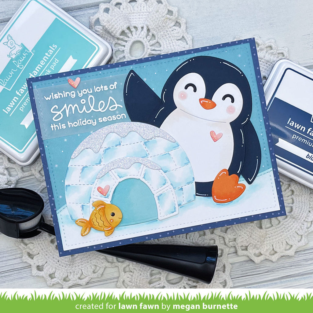 A handmade holiday greeting card featuring a cute penguin, igloo, and goldfish, styled on a white lace doily with Lawn Fawn premium ink pads and a black stamp blade visible around it.